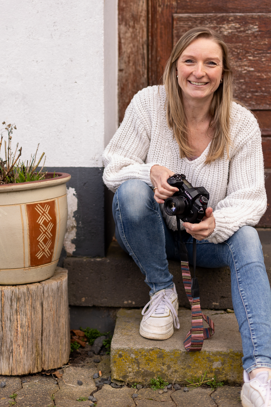 Authentisches Portrait einer Fotografin aus Bonn mit Kamera und freundlichem Lächeln vor einer alten Holztür
