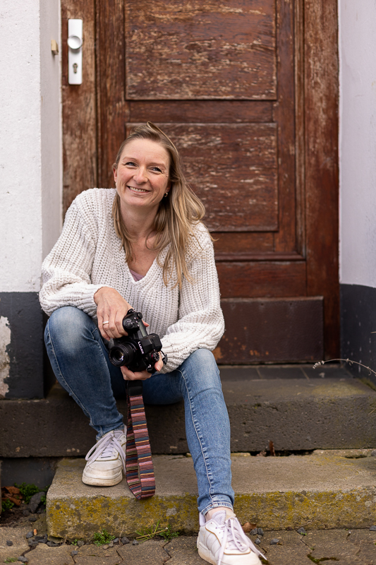 Fotografin aus Bonn mit Kamera lächelt verschmitzt bei einem entspannten Outdoor-Portrait vor einer Holztür