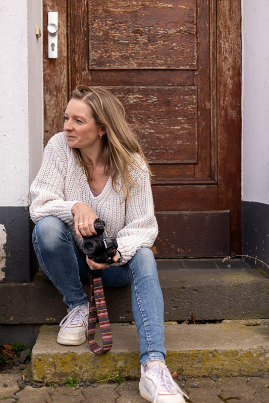 Natürliche Portraitaufnahme einer Fotografin aus Bonn mit Kamera, die schmunzelnd zur Seite schaut
